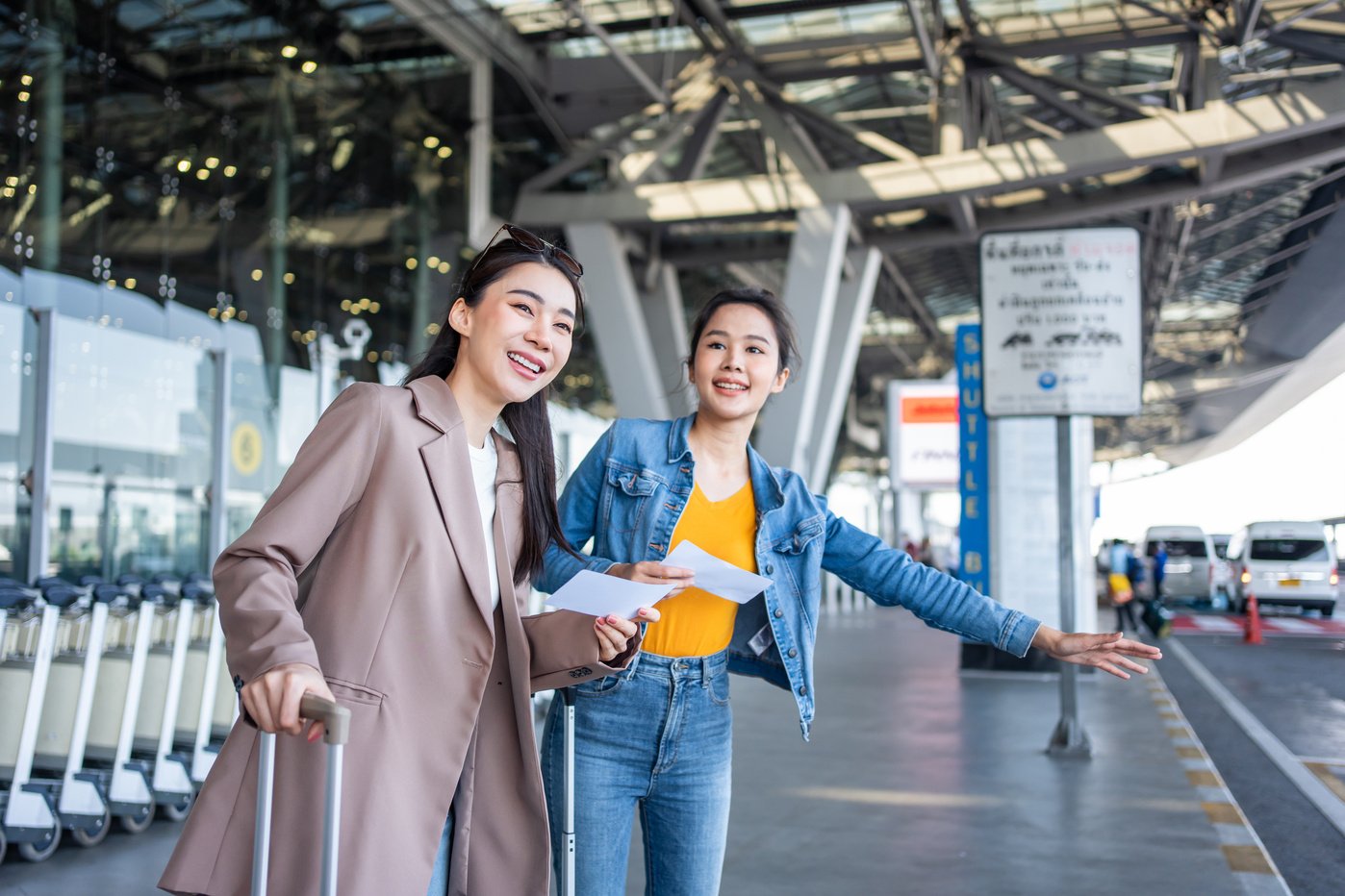 Asian two women passenger waiting a bus after leaving from the airport. Attractive beautiful female tourist friends feeling happy and excited to go travel abroad by airplane for holiday vacation trip.